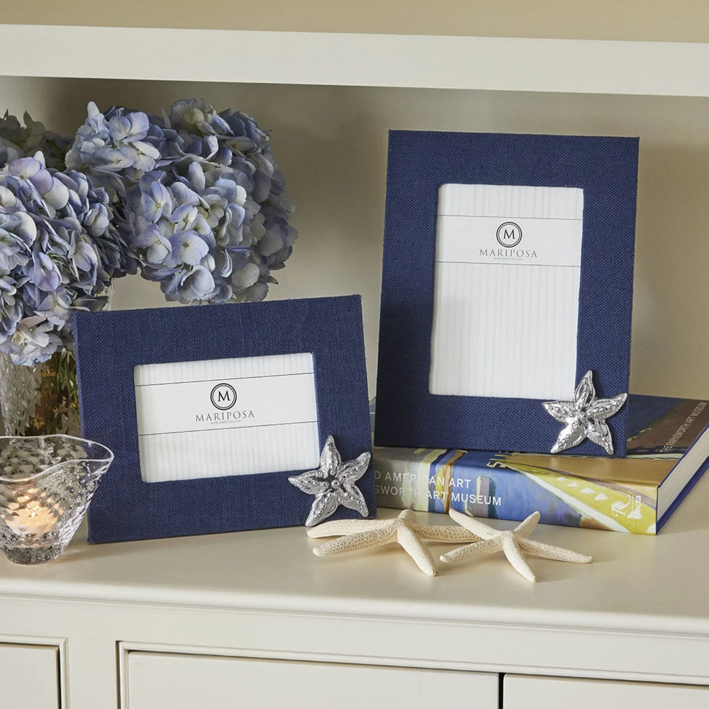 Navy blue 5x7 and 4x6 frame with a silver starfish icon in the bottom left hand corner and 'Mariposa' branding on a white shelf with two white starfish, books and a blue flower with a tealight candle. 