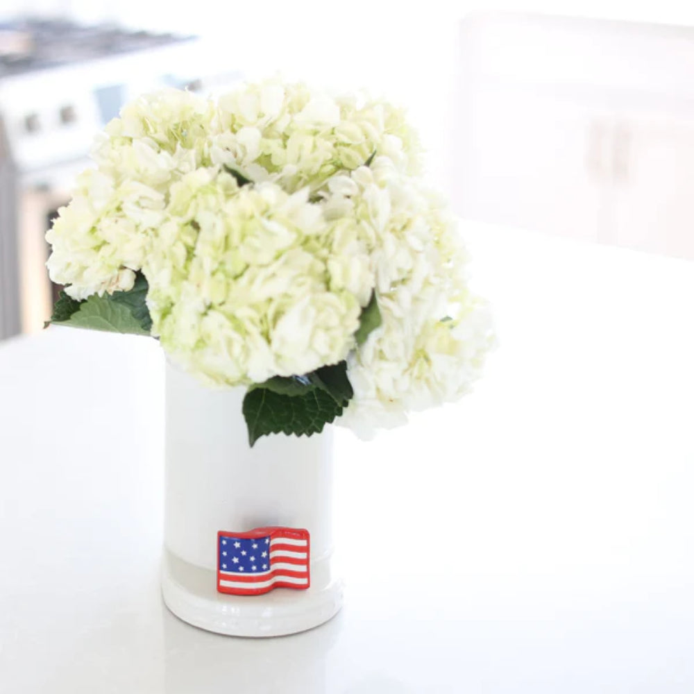 Small rectangular mini decor piece with American flag design on a white vase with white flowers on a white counter top