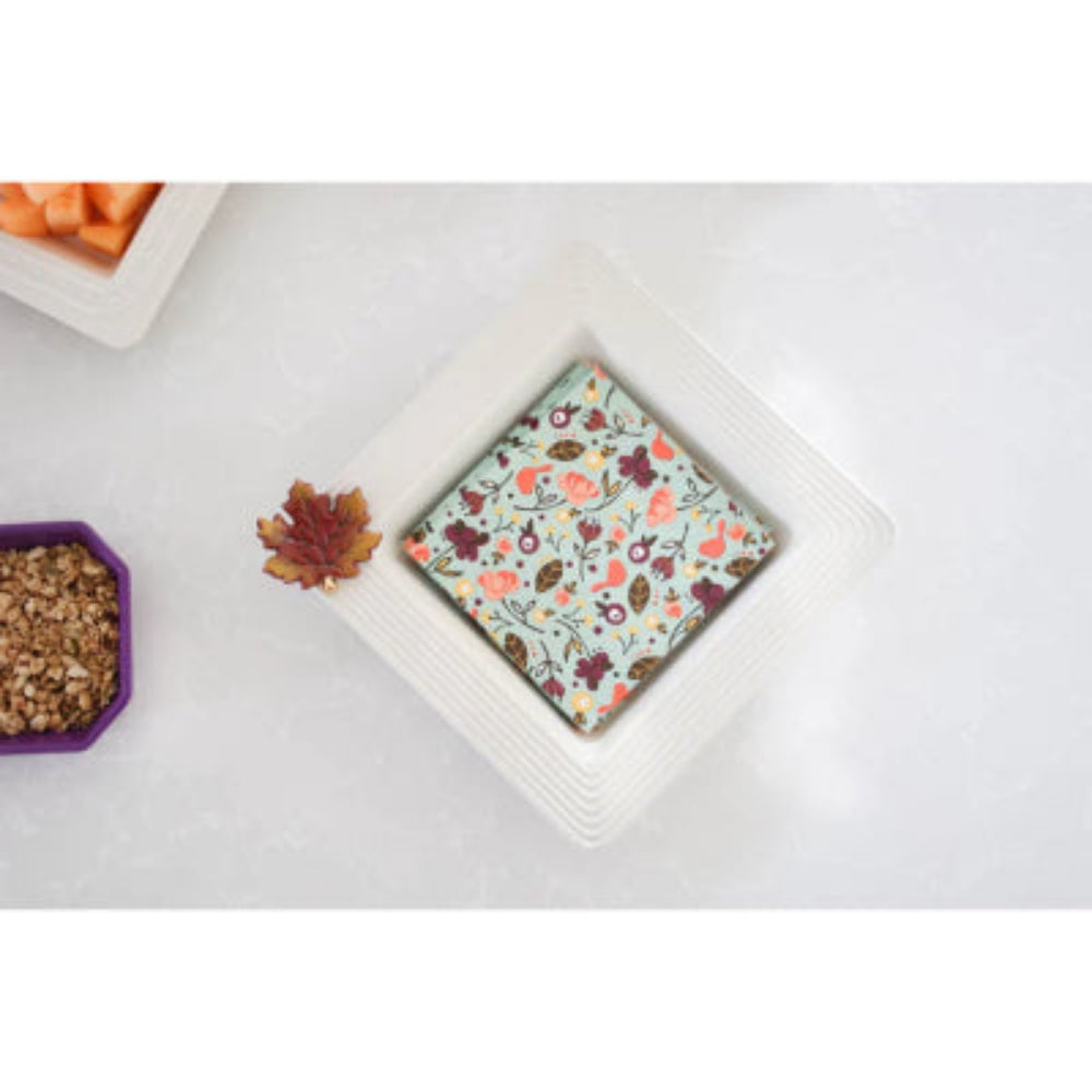 A white square ceramic napkin holder with textured rim on a grey background with a fall leaf decor  and green fall napkins 